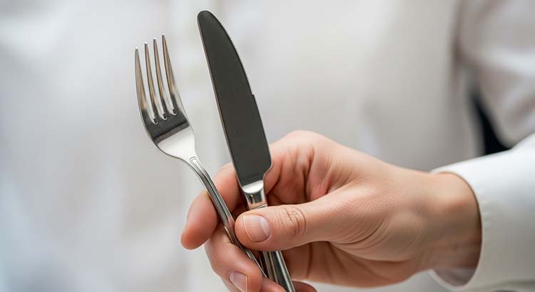 A hand holding a fork and knife, testing its weight and balance against a clean plate