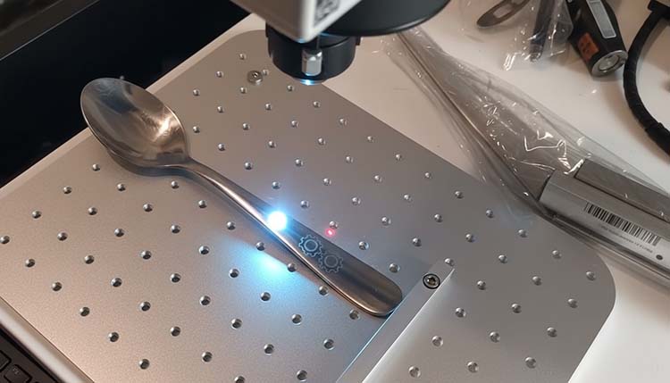 A close-up shot of a fiber laser machine engraving a logo onto the handle of a stainless steel spoon