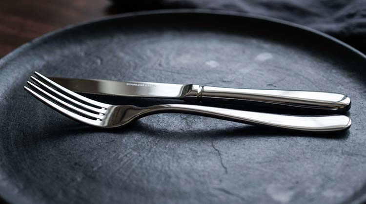 A close-up of a high-quality stainless steel fork and knife on a dark surface