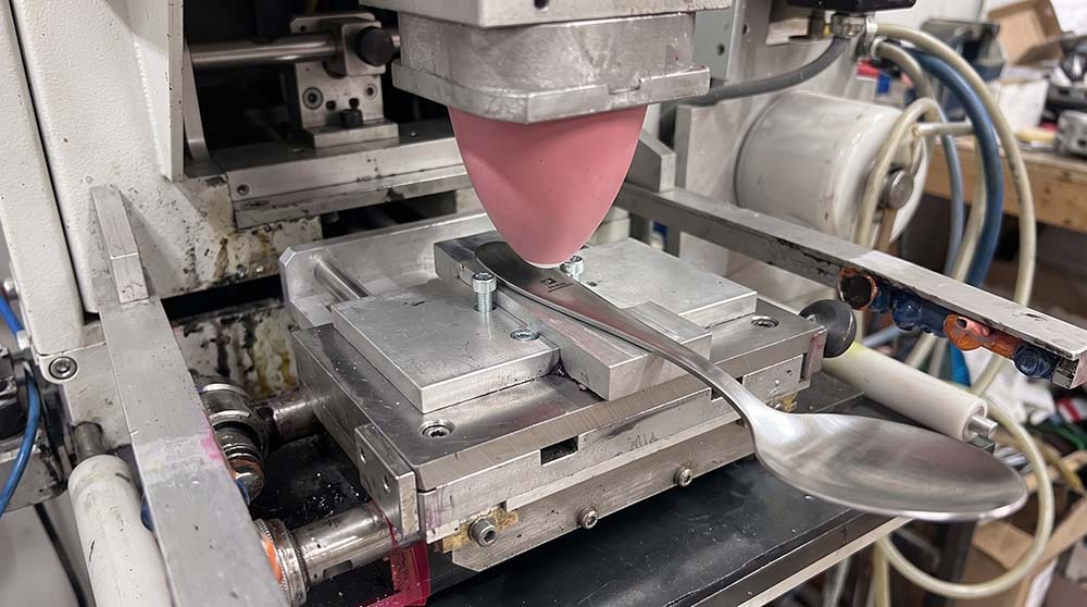 Pad Printing for Stainless Steel Cutlery A close-up of a pad printing machine applying a black logo to a stainless steel spoon handle.